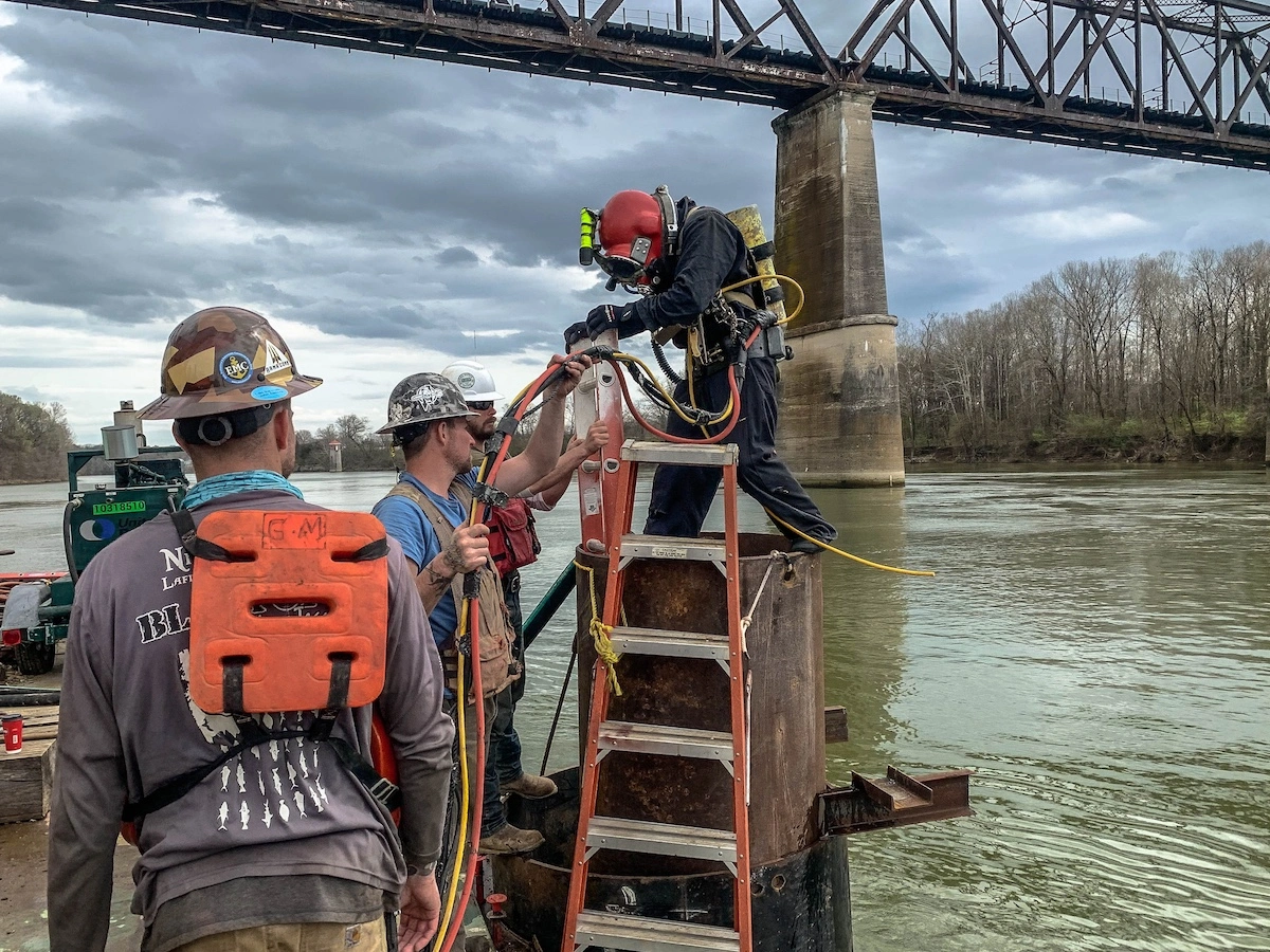 Commercial diving crew supporting a bridge inspection from a ladder beside a Florida waterway