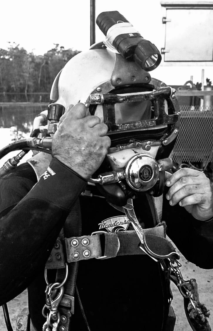 Black-and-white close-up of an ADCI-certified commercial diver fitting his helmet, full harness and gear visible
