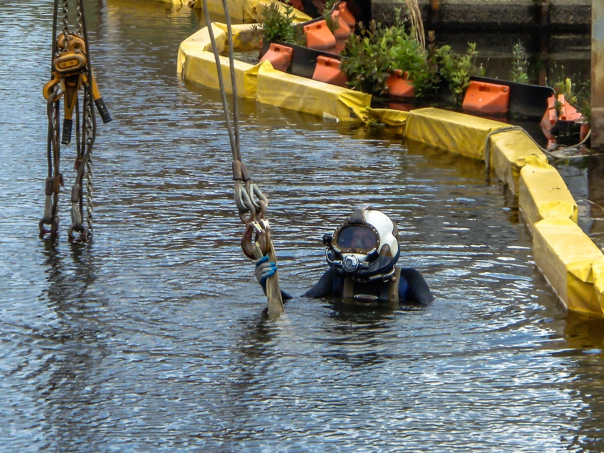 Commercial diver in helmet performing underwater rigging work alongside containment booms in a Florida waterway
